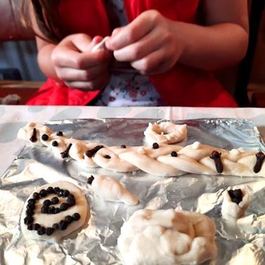 Child's hands working with bread dough and showing a tray of finished pieces decorated by peppercorns.